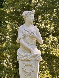 A classical white marble female deity statue wearing a laurel crown and draped robes, set against dappled sunlight through green trees in a European garden. 
