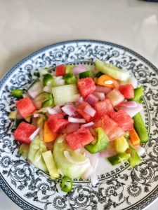 A colorful salad featuring diced watermelon, cucumber, and green bell peppers, mixed with sliced red onions on a decorative black and white plate.