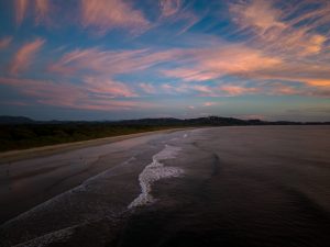 An aerial view of a quiet beach at sunset, with gentle waves rolling toward the shore. The sky is filled with soft pink, orange, and blue clouds, while the coastline curves into the distance beside dark hills.