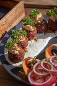 Falafel balls served on herbed white sauce, topped with hummus and fresh parsley, alongside a salad of tomato, red onion, carrot, and cucumber ribbons.
