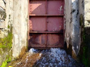 Water gushing beneath a weathered red iron sluice gate set in moss-covered concrete walls — a working irrigation structure in rural Nepal. 