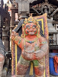 A brightly painted red and green Dvarapala (temple guardian deity) statue holding a sword, with fierce facial expression and ornate jewelry, guarding a Newari temple entrance, Nepal. 