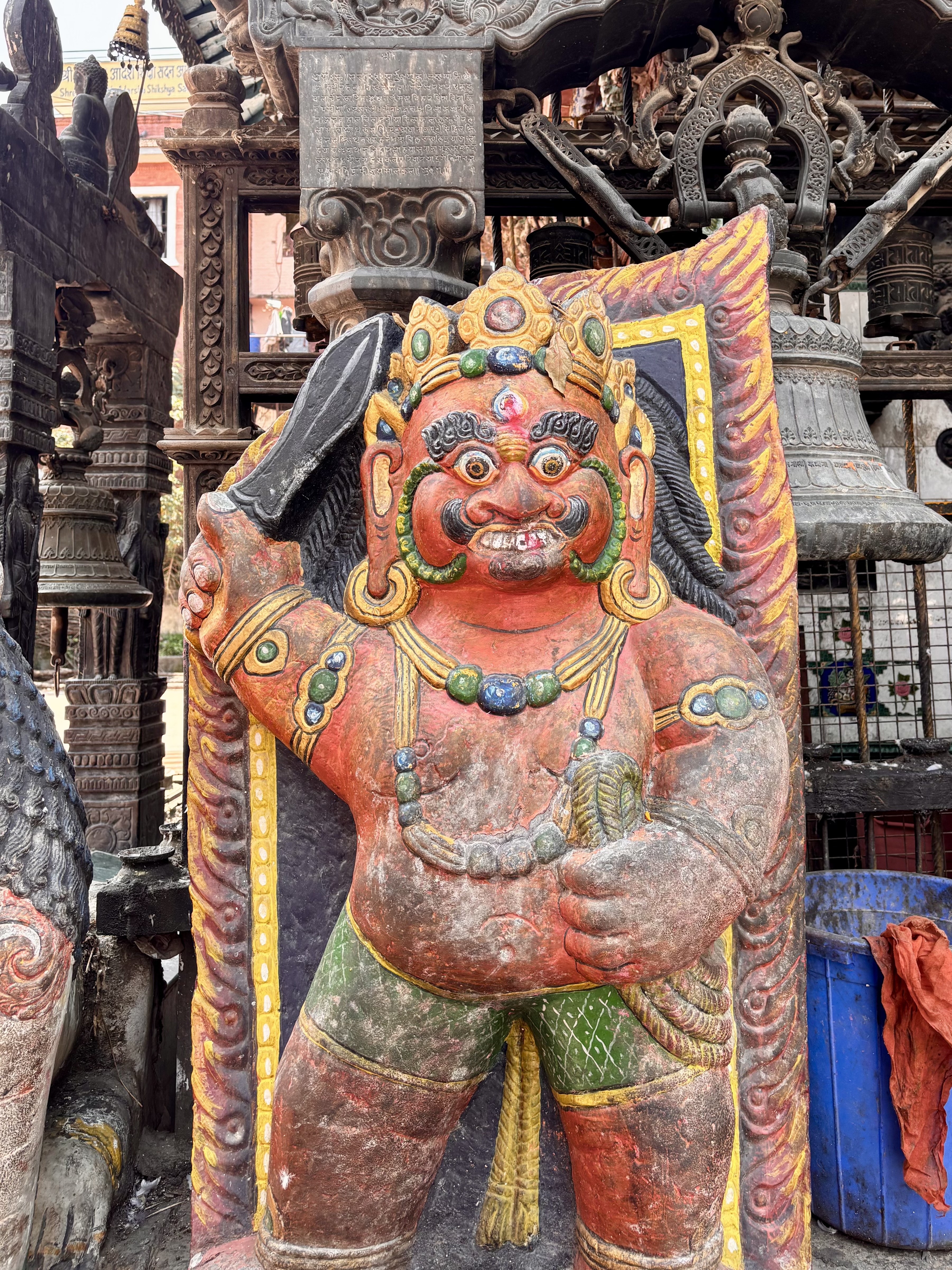 A brightly painted red and green Dvarapala (temple guardian deity) statue holding a sword, with fierce facial expression and ornate jewelry, guarding a Newari temple entrance, Nepal.