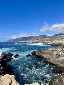A scenic coastal landscape with rocky shores and turquoise waters, where gentle waves crash against the coastline. A small town stretches along the shore, while mountains rise in the distance beneath a clear blue sky dotted with white clouds, creating a peaceful and expansive seaside view.
