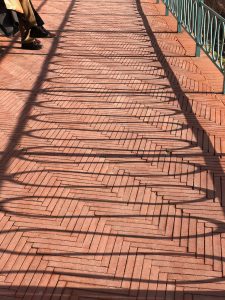 Shadow of a railing projected on a promenade created by bricks that creates a repetitive pattern. On the left side you can see the legs of a person sitting