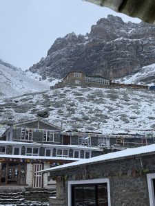 Snow covered stone buildings with colorful prayer flags beneath a towering rocky mountain, conveying a cold, serene atmosphere. Cloudy sky above.