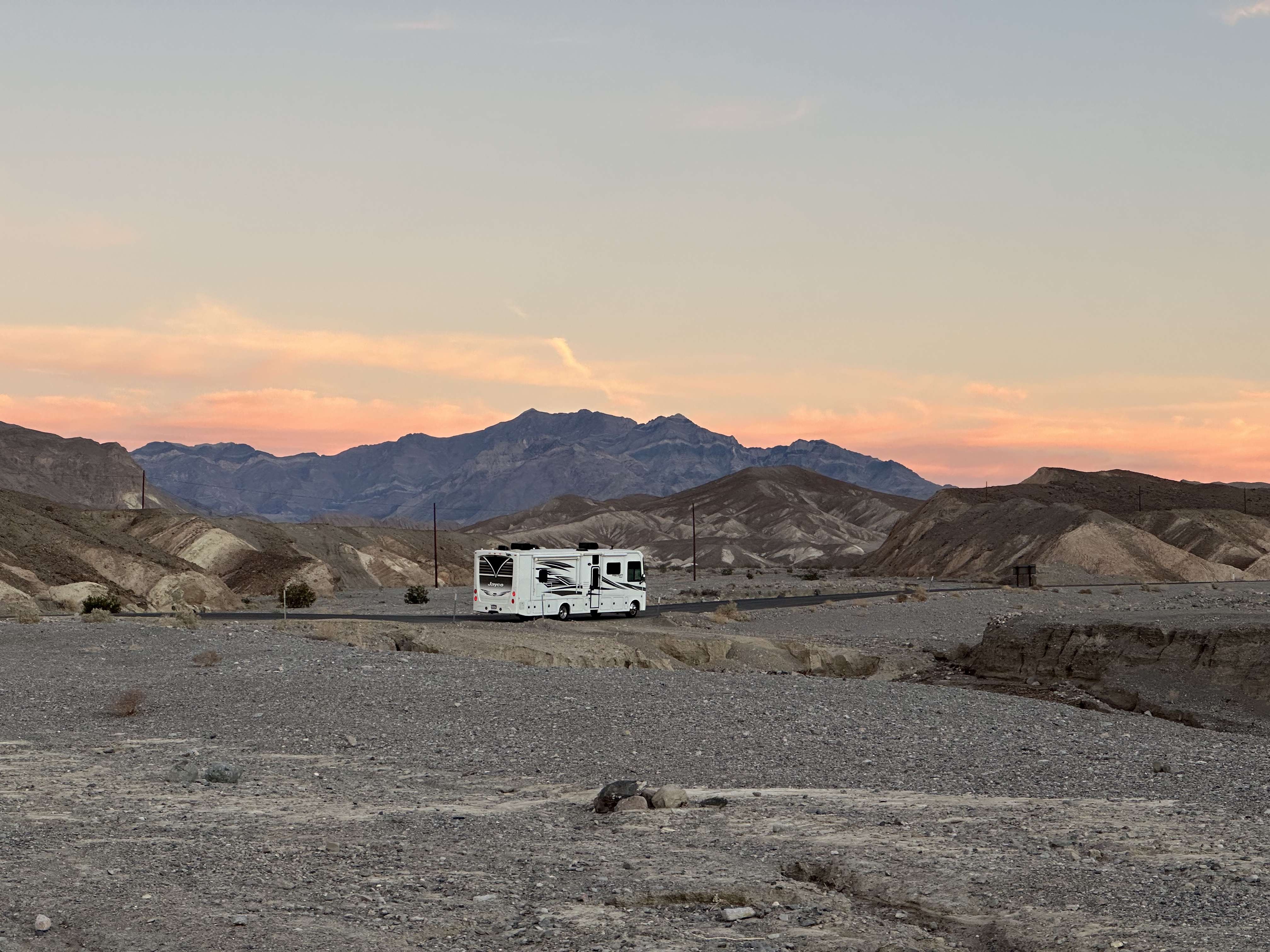 A large white motorhome RV parked on a paved road winding through the vast, rocky badlands of Death Valley National Park. Dramatic, rugged mountain ridges stand sharply against a vibrant pink and orange sunset sky in the background, contrasting with the stark, gray gravel desert floor in the foreground.