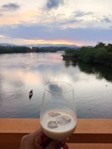 A focused shot of a clear glass filled with an cream drink with ice cubes, resting on an outdoor wooden railing overlooking a wide river. 