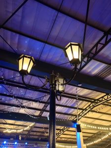 An indoor black lamp post featuring three glowing lantern style light fixtures under a vaulted blue ceiling structure. 
