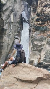 A hiker in a wide-brimmed hat perched on a boulder watching a powerful white waterfall thunder through a narrow sculpted rock gorge. 