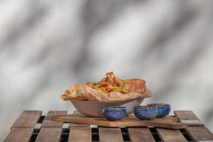 Bowl of crispy potato chips served on brown paper, placed on a wooden board with three small blue dipping bowls on an outdoor table in sunlight.
