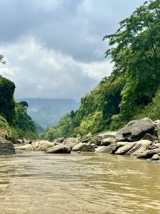 A serene river, Sangu flows between lush green forests and rocky banks under a cloudy sky, with distant mountains in the background, conveying tranquility. 
