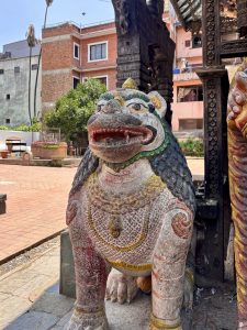 A vibrantly painted polychrome guardian lion (simha) statue with open jaws, colorful jewelry details in red, blue, and yellow, standing at a temple entrance in Kathmandu, Nepal. 