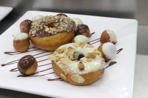 Two donuts with chocolate and white icing topped with nuts, surrounded by mini donut holes with various icing on a white plate. 