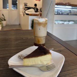 A slice of cheesecake on a white plate with a fork, paired with a layered iced coffee in a glass with a metal straw, set on a wooden table.