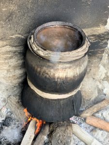 A large dark pot wrapped with cloth sits over an open wood fire, with steam rising from its contents against a rough stone wall, a traditional way of making local alcohol.