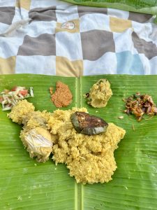 A traditional meal served on a banana leaf, featuring yellow rice topped with eggplant and jackfruit, accompanied by colorful vegetable curries and a spicy paste.
