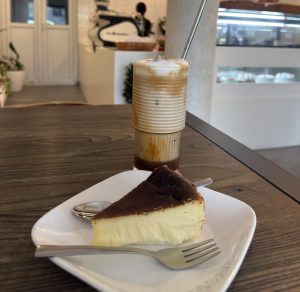 A slice of creamy baked cheesecake served on a white plate with a fork and spoon, paired with a layered iced latte topped with foam on a wooden café table. The softly blurred café interior creates a warm and inviting atmosphere.