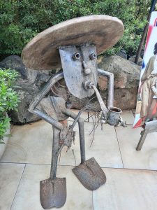 A metal figure sculpture with a wide hat and shovel-shaped feet with a pipe in its mouth.
