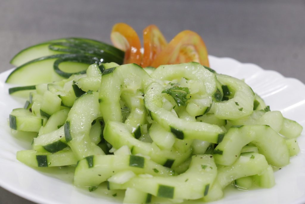 Fresh cucumber salad with herbs on a white plate, garnished with sliced cucumber and tomato in the background.