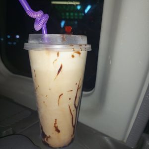 A creamy drink with chocolate drizzle in a clear cup, topped with a lid and a purple curly straw with a red heart.