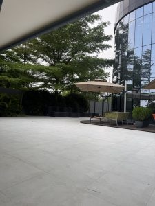Spacious patio with a modern glass building. Large trees and plants surround the area. Beige umbrellas cover outdoor seating,