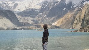 A hooded trekker gazing across a glacial turquoise lake toward towering snow-streaked rock faces — likely Tsho Rolpa or a similar high-altitude glacial lake in Nepal. 