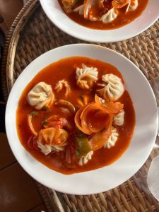 Steaming jhol momo (Nepali dumplings in spiced tomato broth) with sliced carrots and green peppers, served in a white bowl on a wicker tray. 