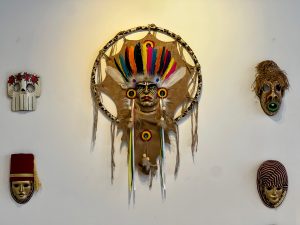 Five decorative cultural masks are displayed on a plain white wall, featuring a large Indigenous-style headdress mask with colorful feathers in the center.
