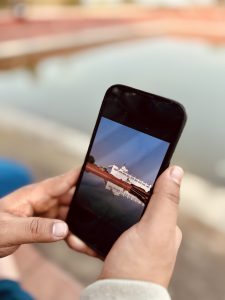 A close-up view of a person's hands holding a black smartphone. The screen displays a clear photo of the white, rectangular Maya Devi Temple building and its distinct geometric reflection mirrored in a still pond. 
