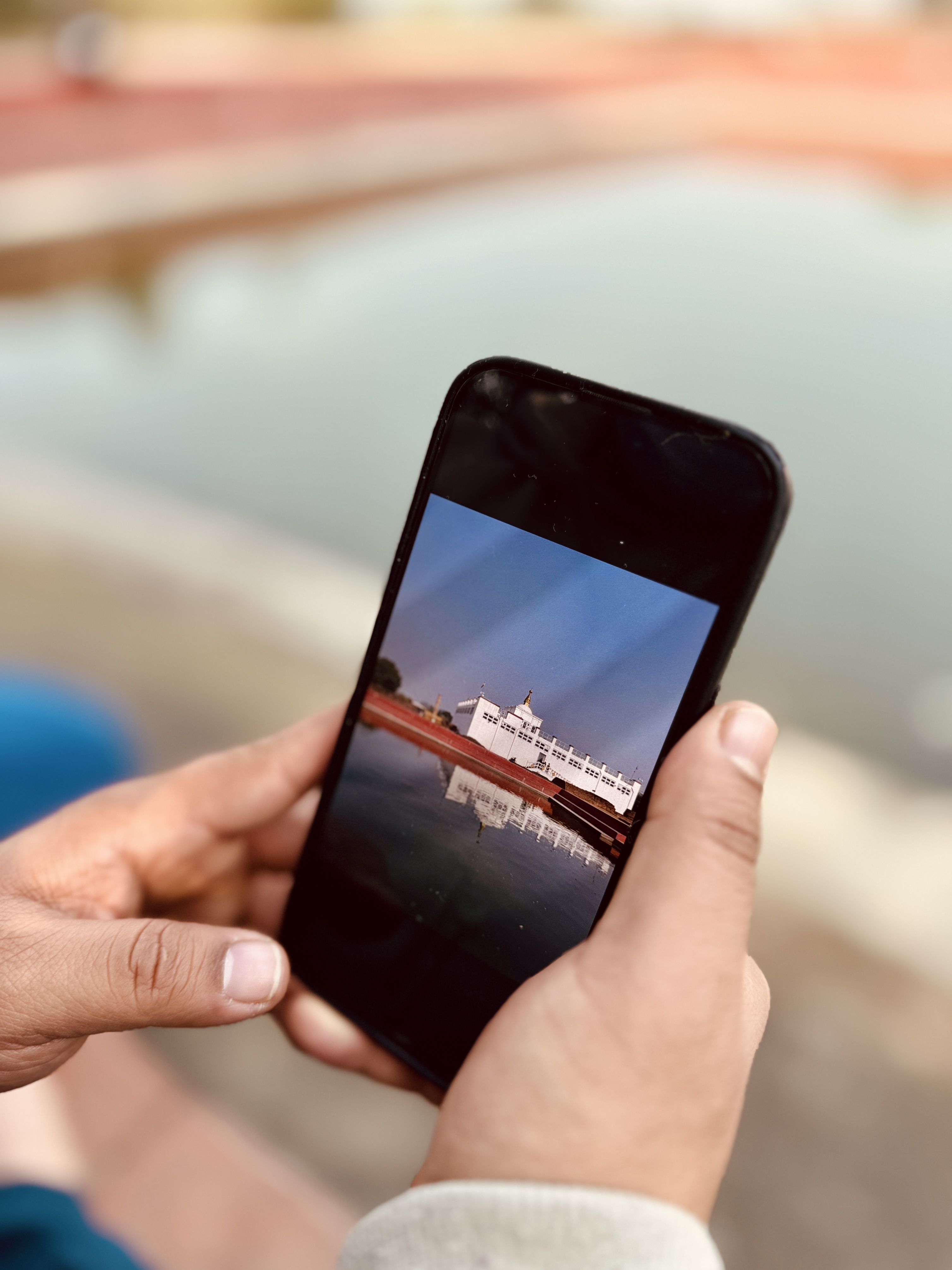 A close-up view of a person's hands holding a black smartphone. The screen displays a clear photo of the white, rectangular Maya Devi Temple building and its distinct geometric reflection mirrored in a still pond.