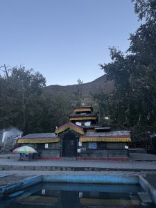 A serene temple with colorful tiers and yellow trim stands against a backdrop of lush trees and a clear sky, reflected in a nearby still pool.
