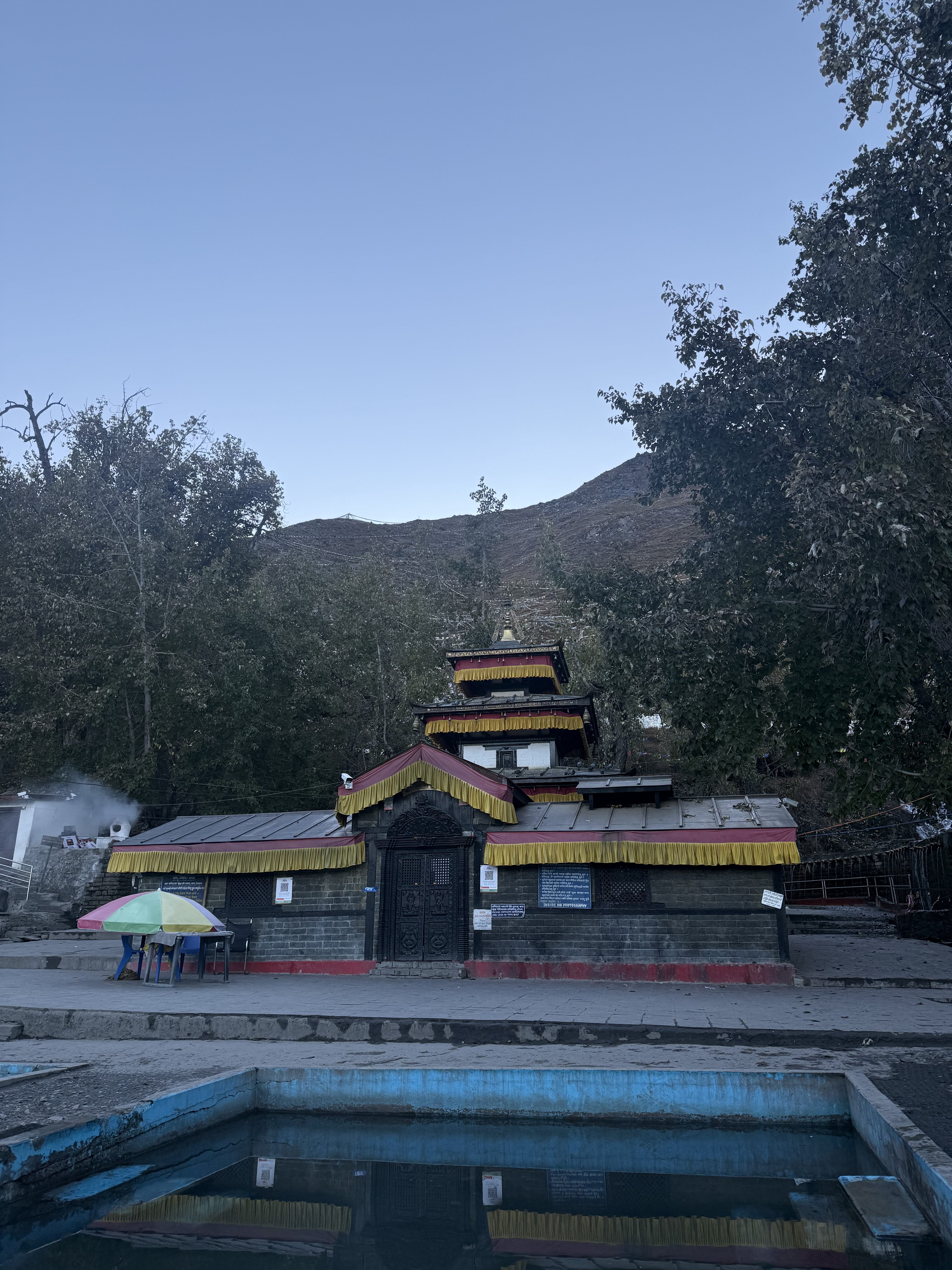 A serene temple with colorful tiers and yellow trim stands against a backdrop of lush trees and a clear sky, reflected in a nearby still pool.
