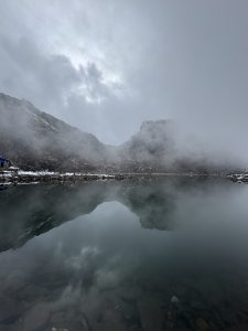 Misty Alpine Lake Mountain Reflection. A vertical, wide-angle shot of a glass-like alpine lake perfectly reflecting the moody landscape above it. In the background, dark, rugged mountain peaks are heavily shrouded in thick, rolling white fog and low-hanging gray storm clouds.
