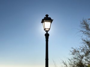 The top of a street lamp that is illuminated by the sun that is just behind with a blue sky in the background. On the right you can see the leaves of a tree.
