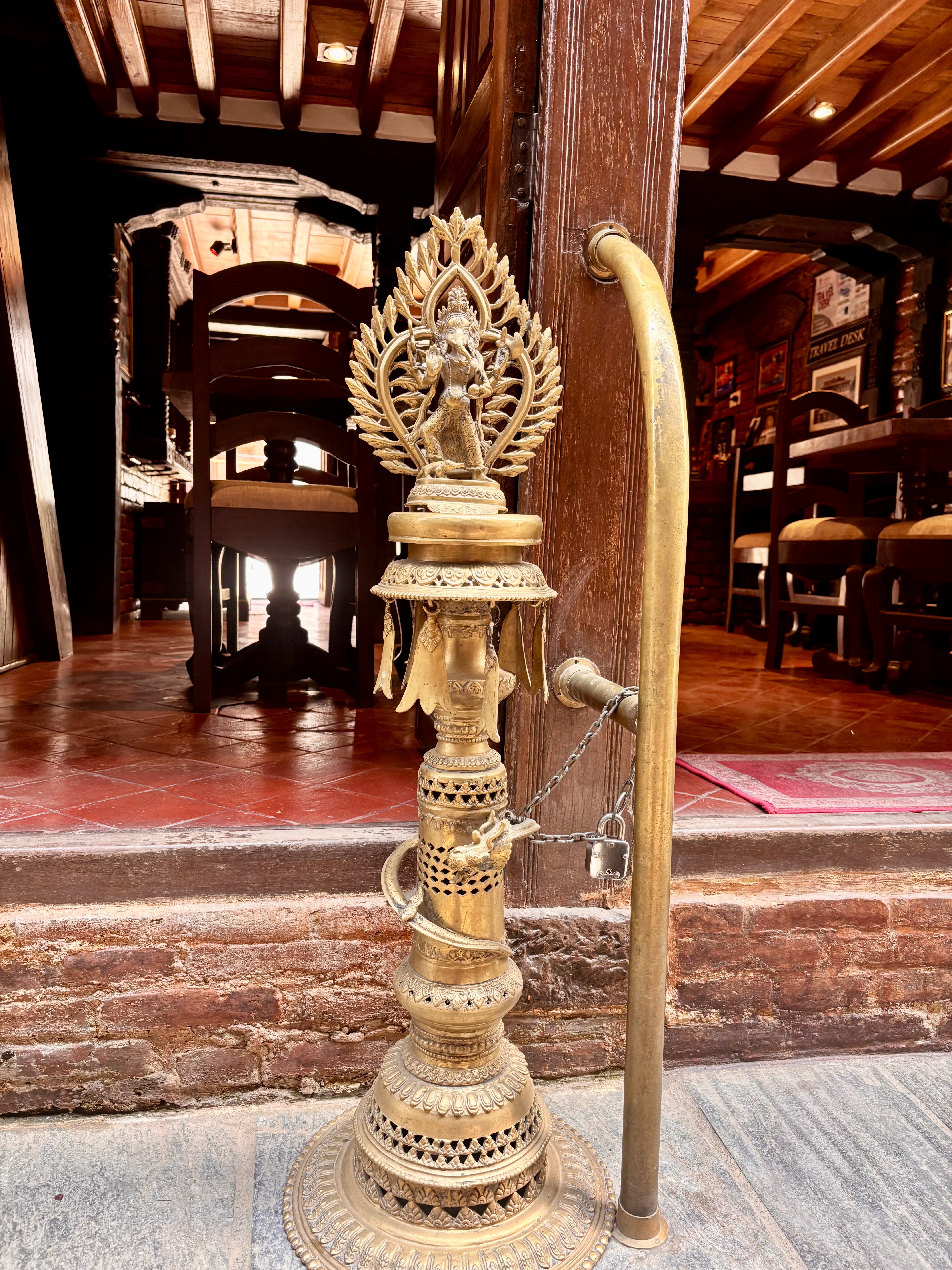 An ornate brass oil lamp pedestal topped with a Ganesh figure surrounded by a flame aureole, chained at the entrance of a traditional Nepali building.