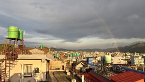Rooftop view of a dense urban skyline dotted with colourful water tanks as a faint rainbow emerges over the hills under a stormy sky.