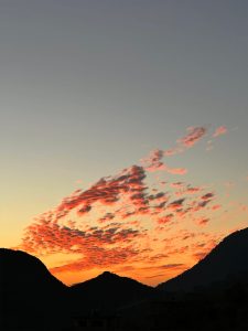 A peaceful sunset paints the sky with shades of blue, orange, pink, and purple, while wispy clouds drift across the horizon. Dark mountain silhouettes frame the scene, and a faint building blends into the foreground, creating a calm and serene end-of-day landscape.