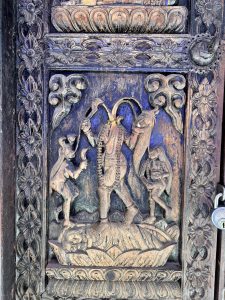 Detailed blue-painted wooden temple door carving depicting a tall figure (possibly Bhairava or Shiva) flanked by smaller figures, standing on a lotus, with padlock visible. 