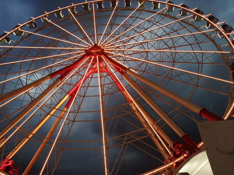 A large, illuminated Ferris wheel structure captured from a low angle against a dark, cloudy evening sky.