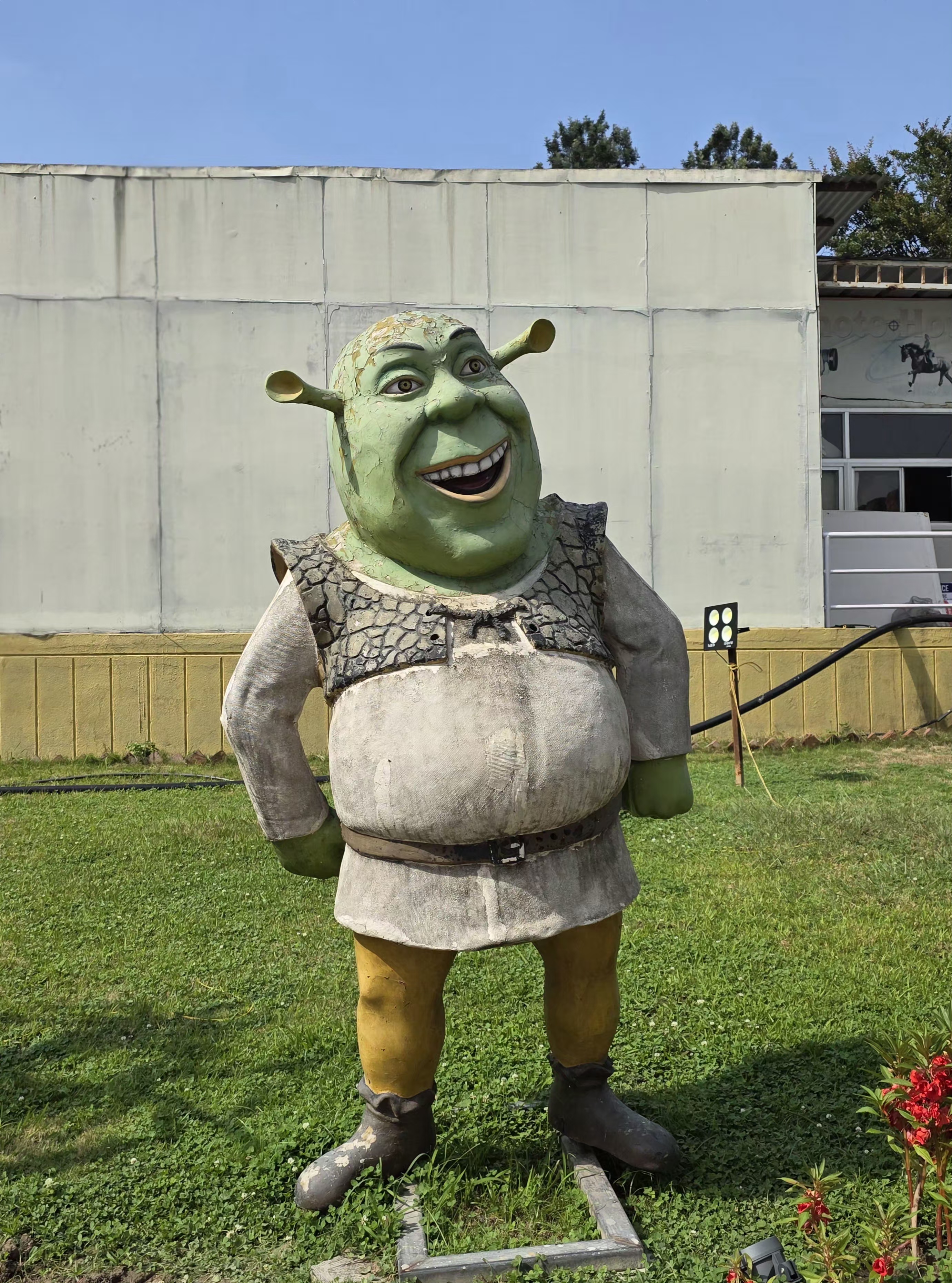 A large statue of a green ogre stands on grass, with hands on hips and a joyful expression. The background features a simple building and clear sky.