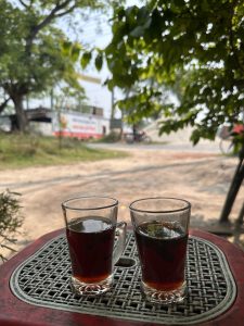 Two glasses of black tea sit on a red, woven-patterned table outdoors. Sunlight filters through lush green leaves, creating a relaxed, serene atmosphere in Kushtia, Bangladesh.
