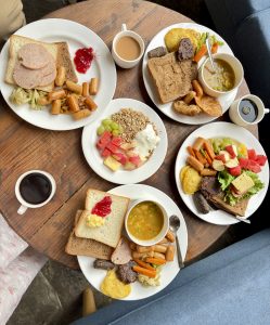 Overhead flatlay of four full English-style breakfast plates with toast, sausages, eggs, vegetables, soup, and fruits, along with granola yogurt bowl and cups of black coffee and milk tea, Nepal.