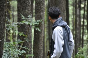 A young man with dark, wavy hair is seen from behind, looking over his shoulder into a dense forest. He wears a grey and white hooded jacket, and a pair of binoculars hangs at his side. He is surrounded by tall, dark tree trunks with rough bark, and scattered green leaves are visible in the foreground.