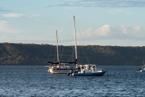 Two boats anchored on calm blue water, including a sailboat with tall masts and a small motorboat in front. A tree-covered hillside and soft clouds appear in the background under warm daylight.