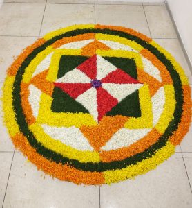 Colorful floral rangoli on tiled floor featuring concentric circles in orange, yellow, green, and white, with a central red and white pinwheel design.
