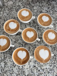 Eight cups of coffee with heart-shaped latte art are arranged on a speckled gray countertop, conveying warmth and hospitality.
