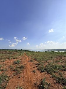 A red dirt path crossing open countryside toward a peaceful lake beneath a bright blue sky with scattered white clouds.