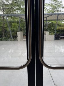 Close-up of metal door handles on glass doors, with raindrops on the glass. Beyond, a patio with geometric planters and lush greenery is visible.