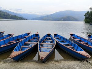 A row of numbered blue wooden rowboats moored side by side on the muddy shore of Begnas Lake, with misty green mountains and cloud-draped hills reflected in the calm water behind them, Pokhara, Nepal. 
